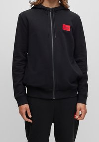 Černá mikina na zip vyrobená z bavlněné směsi, s předním logem v červené barvě, nastavitelnou kapucí se šňůrkou a žebrovanými manžetami a lemem.
