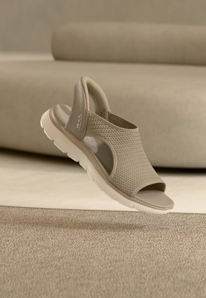 Sandalia beige Skechers sin cordones con parte superior de tela tejida, punta abierta, correa trasera acolchada y suela gruesa de goma blanca sobre suelo alfombrado.