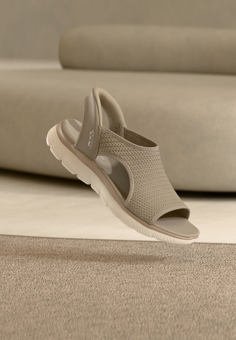 Μπεζ σανδάλι Skechers slip-on με πλεκτό ύφασμα στο πάνω μέρος, ανοιχτά δάχτυλα, επενδεδυμένο λουράκι στο πίσω μέρος και χονδρό λευκό καουτσούκ σόλα πάνω σε στρωμένο χαλί.