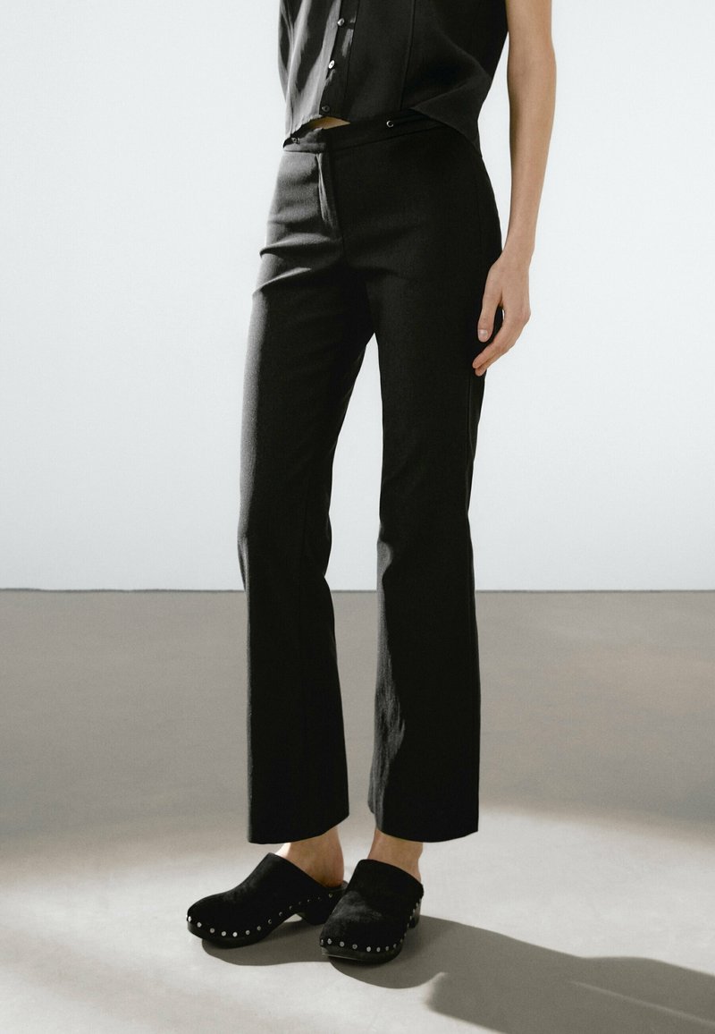 Personne portant un pantalon noir court, un haut noir sans manches et des sabots noirs avec des clous métalliques, se tenant sur un sol uni.