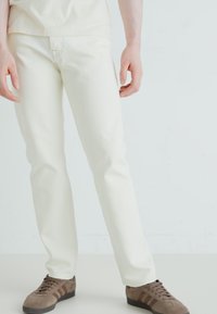 Jeans albi cu croi drept din bumbac, cu o textură netedă și cusături subtile, purtați cu adidași maronii cu dungi.