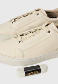 Baskets en cuir beige avec une texture lisse, présentant un design minimaliste, un bout rond et des semelles en caoutchouc plates, accompagnées de lacets assortis.