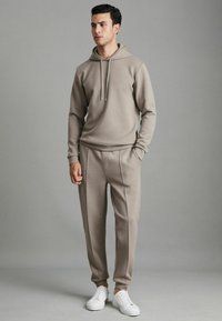 Beige kapucnis pulóver, zsinórral, amelyhez illeszkedő, szűk fazonú melegítőnadrág társul. Fehér sportcipőkkel egészíti ki az öltözéket. Sima szövettextúra.