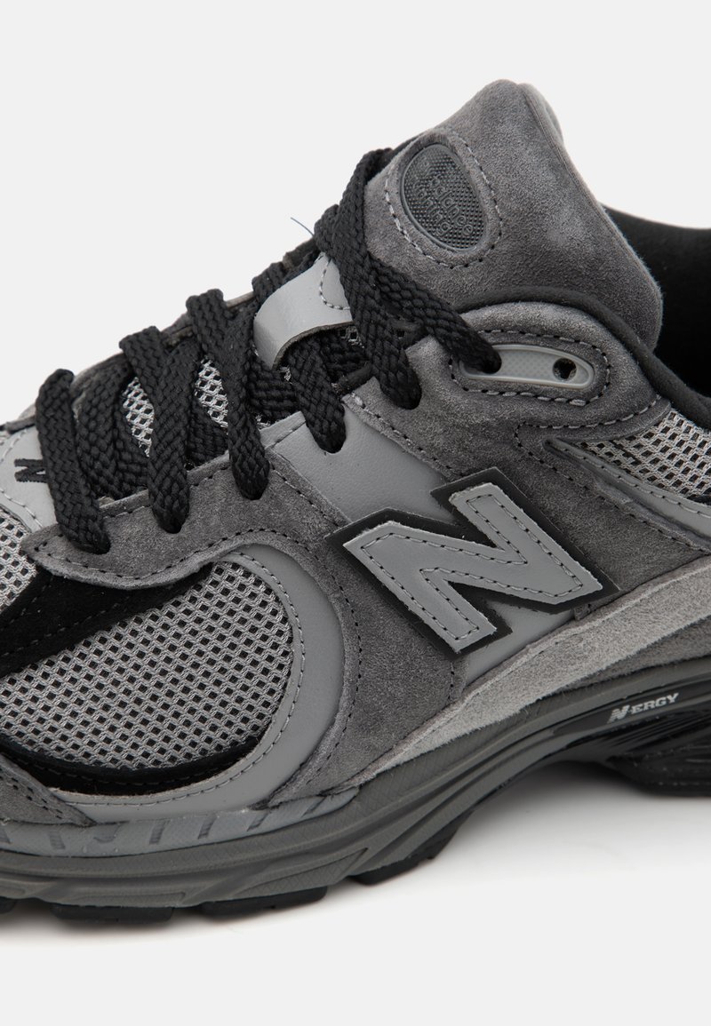 Chaussure de sport grise en maille et daim, avec un col rembourré, des lacets noirs et un logo "N" prominent sur le côté.