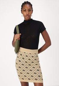 Un col roulé noir à manches courtes associé à une jupe en tricot beige ornée de motifs en diamant noirs et de détails de logo, complétée par un sac vert.