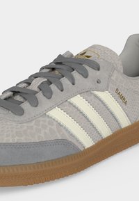 Γκρι αθλητικό παπούτσι Adidas Samba με υφασμάτινο επάνω μέρος, λευκές ρίγες, γκρι κορδόνια, λαστιχένια σόλα και χρυσό λογότυπο στο πλάι και στη γλώσσα.