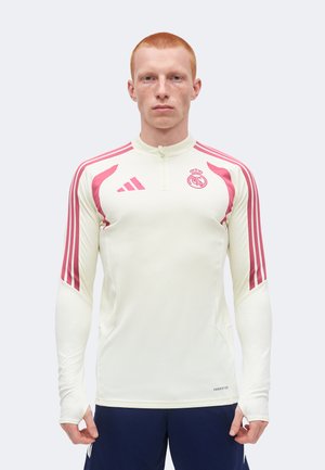 Tânăr cu păr roșu scurt stă cu fața spre înainte, purtând un tricou sport alb cu mâneci lungi cu dungi roz și emblema Real Madrid.