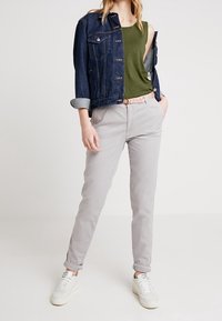 Jeansjacka med upprullade ärmar, olivgrön linne, ljusgrå byxor och vita sneakers visar en avslappnad, lager-på-lager-outfit.