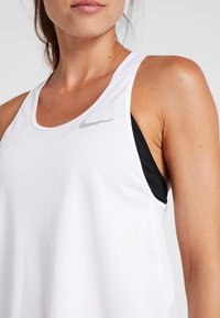 Valkoinen urheilullinen hihaton paita, jossa on pyöreä kaula-aukko. Paidassa on hienovarainen Nike-logo harmaana vasemmassa yläkulmassa ja musta rintaliivi alla. Pintarakennetta on sileä.