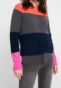 Pletený svetr s horizontálními pruhy v korálové, šedé, námořnické a růžové barvě. Hřejivé náplety a spodní lem, s volným střihem. Nošený přes šedé džíny.