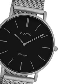 Cadran noir avec des index et des aiguilles en argent, monté sur un bracelet en mailles métalliques argentées. Présente le texte "OOZOO" et "Vintage".
