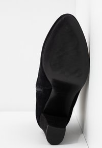 Zapato de tacón de ante negro con punta afilada y suela de goma. Cierre de cremallera lateral y tacón negro texturizado. Líneas limpias y acabado suave.