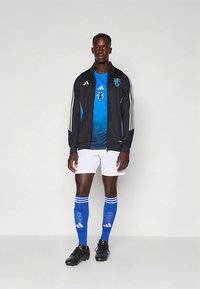 Schwarze Sportjacke mit weißen Streifen, T-Shirt in Blau- und Grauverlauf, weiße Shorts, blaue Socken mit Streifen und schwarze Fußballschuhe.