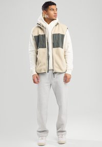 Fleeceweste in Creme und Dunkelgrün mit Reißverschluss und Seitentaschen, über einem weißen Hoodie getragen. Graue Jogginghosen und weiße Sneaker runden den Look ab.