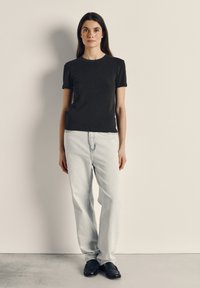 Moteris su ilgais tamsiais plaukais stovinti, apsirengusi juodus trumpomis rankovėmis marškinėlius, šviesiai mėlynas džinsas ir juodus loafers, stovinti priešais paprastą foną.