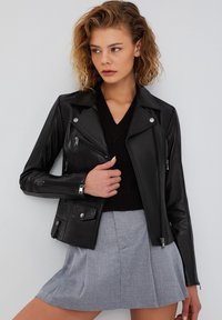 Giacca da biker in pelle nera con tasche con zip, dettagli in metallo argentato e un design attillato. Abbinata con un maglione scuro e una gonna grigia.