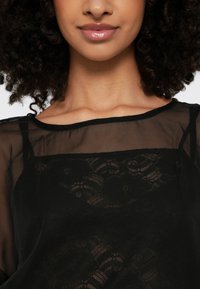 Schwarze Bluse mit durchscheinender oberer Schicht, Spitzenverzierung und weitem Ausschnitt. Weiche Textur mit eleganten dekorativen Mustern.