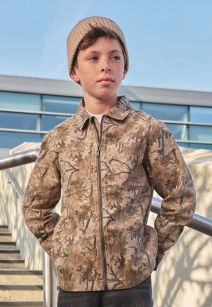 Jonge jongen met sproeten, die een beige muts en een jas met camouflagepatroon draagt, staat buiten bij metalen leuningen en trappen.