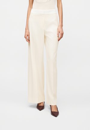 Frau trägt hoch taillierte, weit geschnittene cremefarbene Hose und braune spitze Absätze, steht vor einem schlichten weißen Hintergrund.