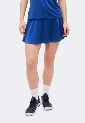 Személy kék teniszszoknyát és felsőt visel, fehér sportzoknit és fekete-fehér sportcipőt, fehér háttér előtt áll.