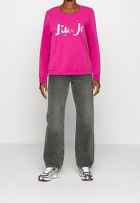 Růžová mikina s dlouhými rukávy a kulatým výstřihem, zdobená bílým logem s ozdobami. Nošena s šedými širokými džíny a teniskami.