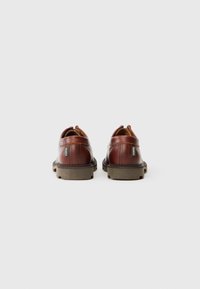 Zapatos de cuero marrón con un acabado suave, con la parte trasera redondeada y suelas de goma texturizadas. Presentan una etiqueta en el lateral y costuras en contraste.