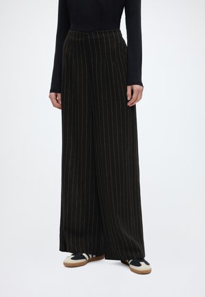 Fekete, széles szárú csíkos nadrágot és fekete hosszú ujjú felsőt viselő személy, fehér és fekete tornacipőt viselve, állva.