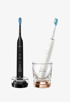 Zwei Philips Sonicare elektrische Zahnbürsten, eine schwarze auf einer Ladestation, eine weiße in einem klaren Glas mit kupfernem Fuß, beide mit blauen Bürstenköpfen.