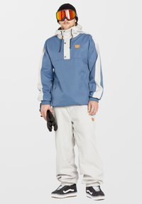 Veste de neige bleue et blanche avec fermeture éclair et boutons, pantalon blanc et baskets noires. Les accessoires incluent des gants noirs et des lunettes de ski avec verres miroités.