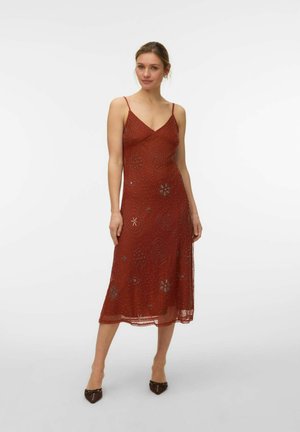 Moteris stovinti dėvinti rūdžių spalvos, be rankovių, midi ilgio suknelę su karoliukais dekoruotais gėlių raštais ir dėvinti rudus aštriais smailėmis aukštakulnius, ant vientisos baltos fono.