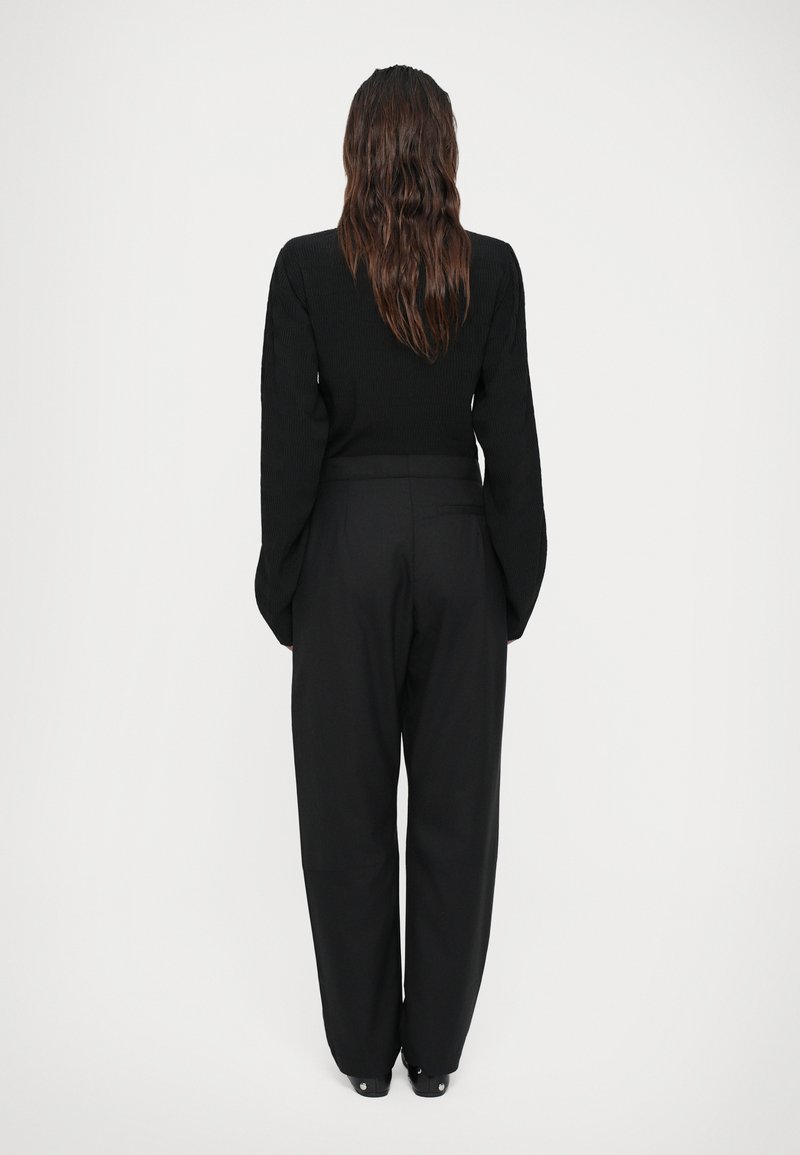 Femme avec de longs cheveux bruns portant un haut à manches longues noir et un pantalon large taille haute noir, se tenant de dos sur un fond blanc.