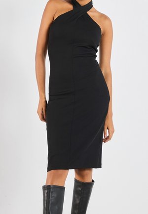 Frau trägt ein ärmelloses, schwarzfarbenes, knieumspielendes Kleid mit Neckholder-Ausschnitt, kombiniert mit schwarzen kniehohen Stiefeln vor einfarbigem Hintergrund.