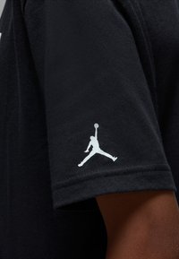 T-shirt noir à texture lisse ; présente un logo Jordan blanc sur la manche gauche. Design simple, manches courtes, et col rond.