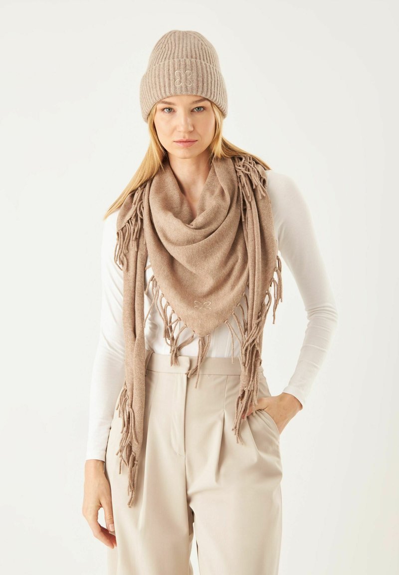 Bonnet en tricot beige et écharpe oversize assortie avec des franges. L'écharpe est triangulaire, de texture douce, portée sur une chemise blanche à manches longues et un pantalon beige.