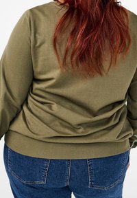 Sudadera verde oliva con un ajuste relajado y dobladillo acanalado, combinada con jeans azules. La tela parece suave y presenta pliegues visibles en la parte posterior.