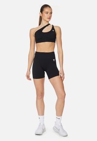 Črn športni modrček z dizajnom enega ramena in izrezom, v kombinaciji z visokoraslimi črnimi kratkimi hlačami. Obe podjetji sta izdelani iz gladkega, raztegljivega materiala.
