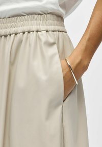 Gros plan d'une main avec un bracelet en argent reposant dans la poche d'une jupe beige à taille élastique sur fond blanc.