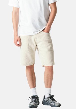 Férfi bézs térdig érő rövidnadrágban, fehér pólóban, fehér bokazokniban és szürke sportcipőben, fehér háttér előtt állva.