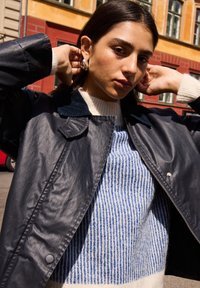 Marineblaue Lederjacke mit einem Cordkragen über einem creme- und blau gestreiften Pullover. Glatte Textur und Details des Reißverschlusses sichtbar.