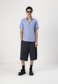 Chemise à manches courtes bleu clair avec un pocket, associée à un short ample foncé et des bottes noires à lacets épaisses. Lunettes de soleil noires portées.