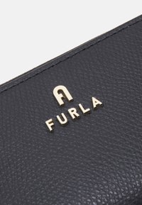 Crni kožni novčanik s teksturom koji ima zlatno urezani 'FURLA' logo. Površina prikazuje fini uzorak zrna, dodajući suptilnu eleganciju.