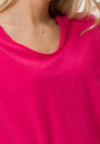 Světlý růžový top s krátkým rukávem a širokým výstřihem, vyrobený z měkké, lehké tkaniny; hladký povrch s jemným leskem.