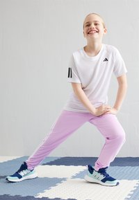 Camiseta deportiva blanca con rayas negras, pantalones de chándal morados claros con rayas laterales blancas y zapatillas verde azulado con suela negra.