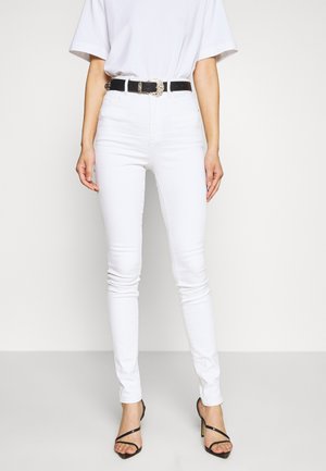 Femme portant un jean skinny blanc taille haute, ceinture noire avec boucle décorative, t-shirt blanc et sandales noires à talons, debout.