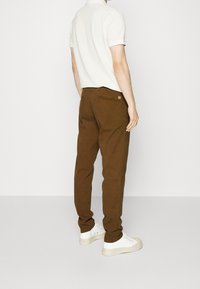 Mężczyzna w brązowych prostych spodniach, białej koszulce polo z krótkim rękawem i białych trampkach, stojący na tle jasnego, jednolitego tła.