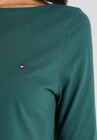 Groene lange mouwen katoenen shirt met een platte halslijn, voorzien van een klein logo-patch in rood, wit en blauw op de borst. Soepele textuur.