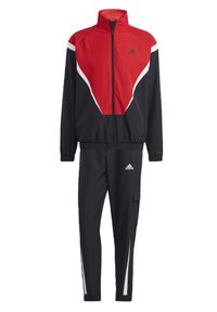 Tuta rossa e nera con giacca con zip, colletto alto e dettagli accentati bianchi. I pantaloni presentano strisce laterali e una tasca utilitaria.