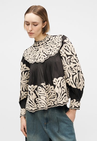 Femme portant un chemisier noir avec une grande broderie florale beige sur les manches, le col et l'ourlet, associée à un jean bleu, regardant vers le bas.