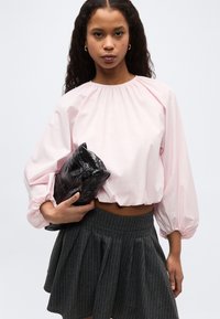 Νεαρή γυναίκα με μακριά σγουρά μαλλιά φοράει ένα ανοιχτό ροζ πουκάμισο με φουσκωτά μανίκια και μια σκουρόχρωμη πλισέ φούστα με ρίγες, κρατώντας μια μαύρη τσάντα clutch.