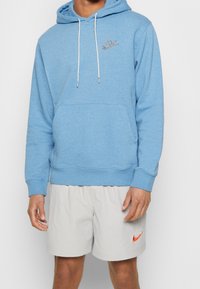 Ljusblå fleece hoodie med en framsida ficka och dragsko i huvan. Har en grå Nike-logotyp på bröstet. Matchas med grå rutiga shorts.